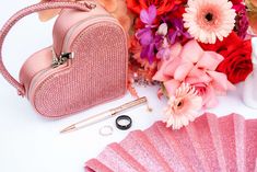 a pink purse next to some flowers and a fan on a white table with other items