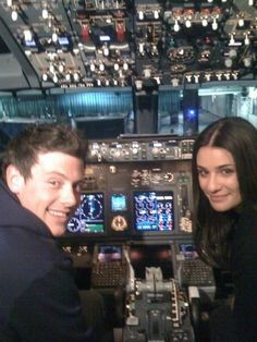 a man and woman sitting in the cockpit of an airplane looking at the camera smiling