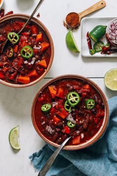 two bowls filled with chili and vegetables on top of a white table next to sliced limes