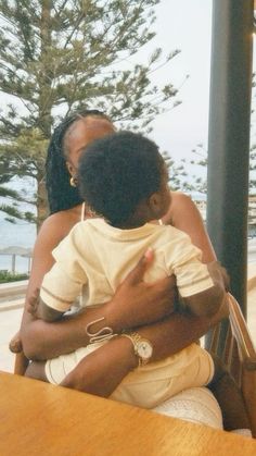 a woman holding a child sitting at a table in front of a window with the ocean behind her