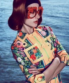 a woman with red hair and glasses on her face is standing near the water wearing an orange floral shirt