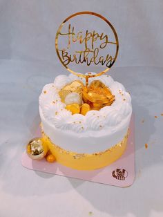 a white cake with gold decorations and happy birthday written on top, sitting on a pink place mat