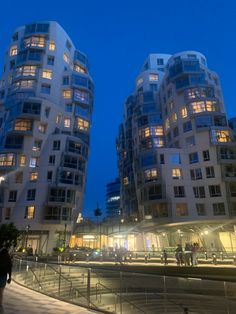 two tall buildings are lit up in the night time, with people walking around them