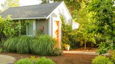 a small gray house surrounded by trees and plants in front of the door is a gravel path leading up to it