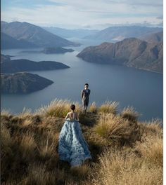 a man and woman standing on top of a grass covered hill next to a body of water