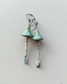 two small green and blue earrings hanging from hooks on a white surface with one dangling off the hook