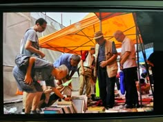 several men are looking at books under an orange umbrella and some people in the background