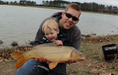 a man holding a fish while sitting next to a little boy in front of a lake