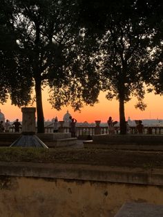 the sun is setting behind some trees and people are sitting on benches in front of them