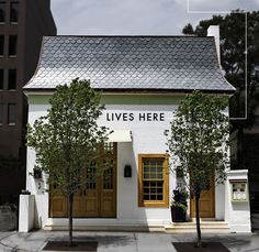 a small white house with two trees in front of it and buildings on the other side