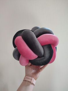 a hand holding up a pink, black and grey ring plush toy in front of a white wall