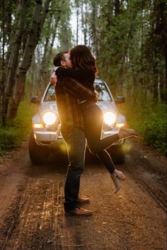 a man and woman kissing in front of a car on a dirt road surrounded by trees