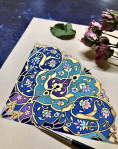 a blue and gold design on a piece of paper next to some purple flowers with green leaves