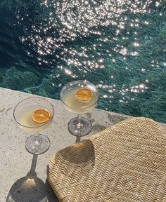 two glasses of wine sitting on top of a table next to a swimming pool with sun reflecting off the water