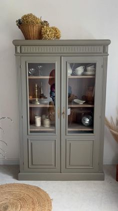 a gray china cabinet with glass doors and baskets on the top, next to a rug