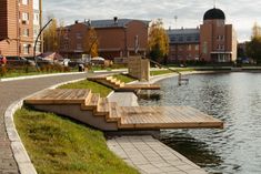 a wooden dock sitting next to a lake in front of tall brick buildings and green grass