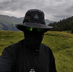 a man wearing a black hat and green eyes is standing in a field with mountains behind him