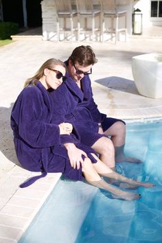 a man and woman in robes sitting on the edge of a swimming pool next to each other