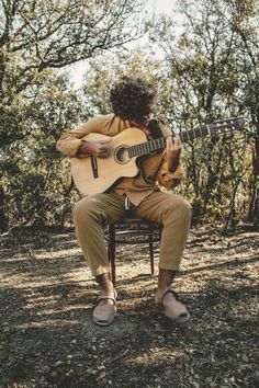 a man sitting on a chair playing an acoustic guitar in the woods with trees behind him