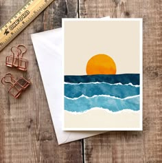 a card with an orange sun over the ocean on it next to some paper clips