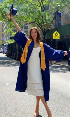 a woman in white dress and blue graduation robe holding her arms up with both hands