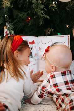 two young children are sitting in front of a christmas tree and looking at an open book