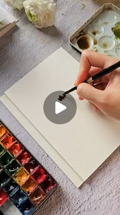 a person holding a pen and writing on a piece of paper with watercolors in the background