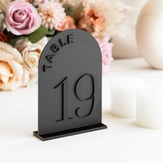 a black table number stands next to some flowers and candles on a white surface with pink roses in the background