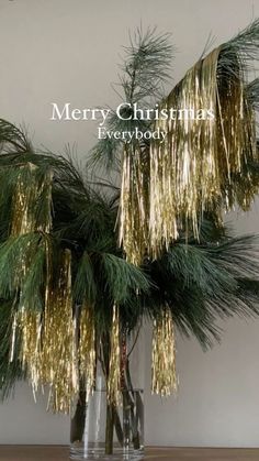 two clear vases filled with pine branches and gold tinsel on top of a wooden table