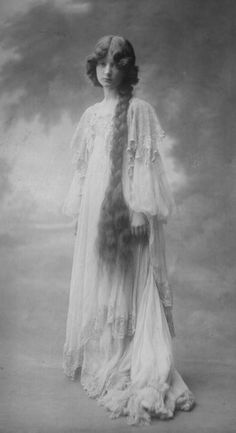 an old photo of a woman with long hair wearing a white dress and standing in front of a cloudy sky