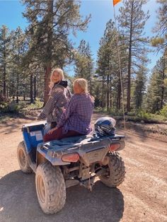 two people riding an atv on a dirt road in the woods with pine trees behind them