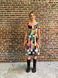 a woman standing in front of a brick wall wearing a multicolored dress and black boots