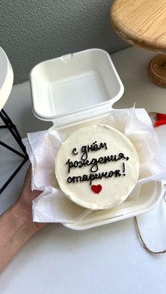 a cake with writing on it sitting in a white box next to a hand holding a plastic container