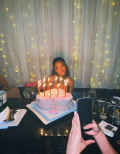 a woman sitting in front of a birthday cake with lit candles on it and her cell phone