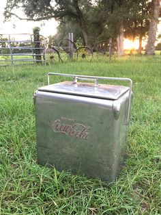 an old coca - cola cooler sitting in the grass near a fence and some trees