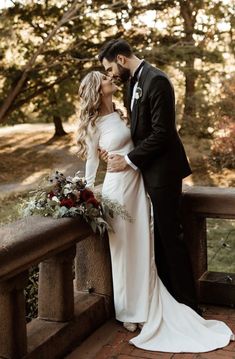 a bride and groom standing on a bridge in front of some trees with their arms around each other