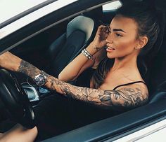 a woman sitting in a car with tattoos on her arms and arm, holding the steering wheel