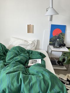 an unmade bed with green comforter in a white room next to a lamp