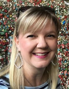 a woman with blonde hair and large earrings smiles for the camera in front of a colorful wall