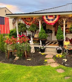 a porch covered in flowers and potted plants with an american flag hanging from the roof