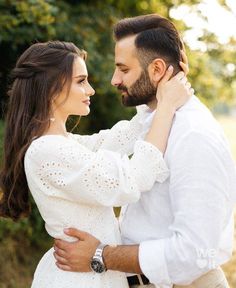 a man and woman standing next to each other in front of trees with their arms around each other