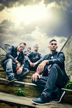 three men are sitting on the steps in front of a brick wall and cloudy sky