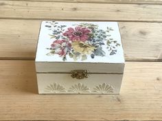 a small white box with flowers painted on the lid sitting on a wooden floor next to a pair of scissors