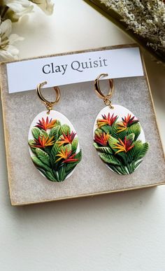 a pair of earrings sitting on top of a white box next to flowers and leaves