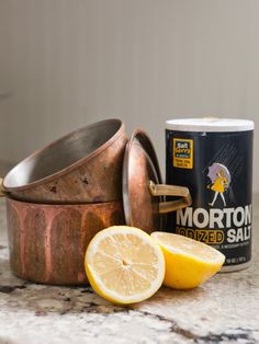 a can of lemon next to some sliced lemons on a marble counter top with a copper pot