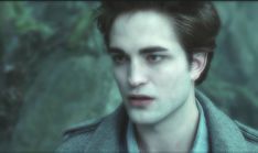 a young man with dark hair and blue eyes looks into the camera while standing in front of a forest