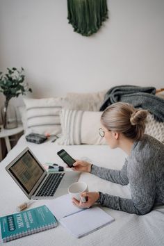 a woman sitting on her bed looking at her laptop and holding a cup of coffee