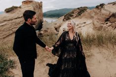 a man and woman holding hands while standing on sand dunes near the ocean with cliffs in the background