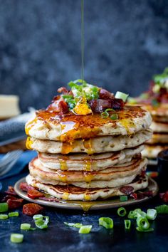 a stack of pancakes with syrup being poured on top and topped with bacon, cheddar cheese, and green onions