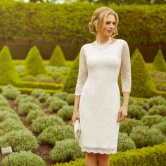 a woman wearing a white dress and heels standing in front of some bushes with her hand on her hip